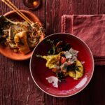 A plate of pan-seared trout with noodles and vegetables next to a red bowl of creamy polenta with wild mushrooms and shaved parmesan, all on a weathered red wooden surface with a red napkin.