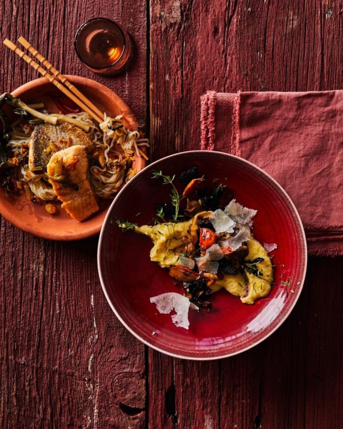 Pan Sauteed Trout with Polenta and Wild Mushroom TABLE Magazine A plate of pan-seared trout with noodles and vegetables next to a red bowl of creamy polenta with wild mushrooms and shaved parmesan, all on a weathered red wooden surface with a red napkin.