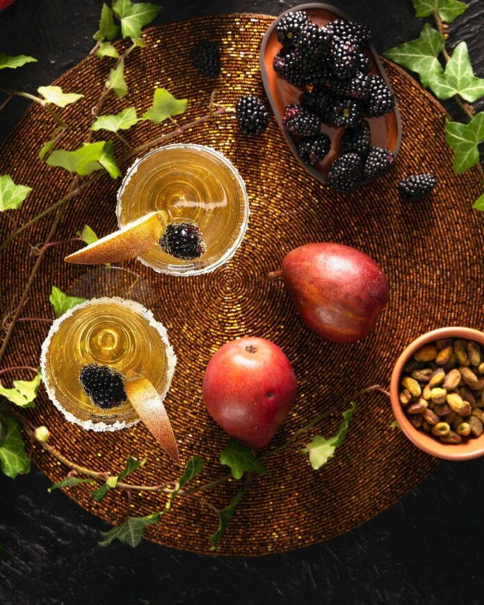 Pear Cider and Gin Bellini A pair of pear cider and gin bellinis in sugar-rimmed glasses, garnished with blackberries and pear slices, on a textured placemat.
