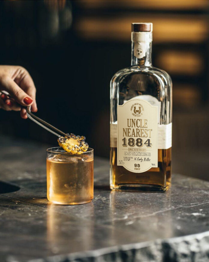 A person places a garnish on a cocktail glass as a bottle of whiskey sits behind it on a marble counter.