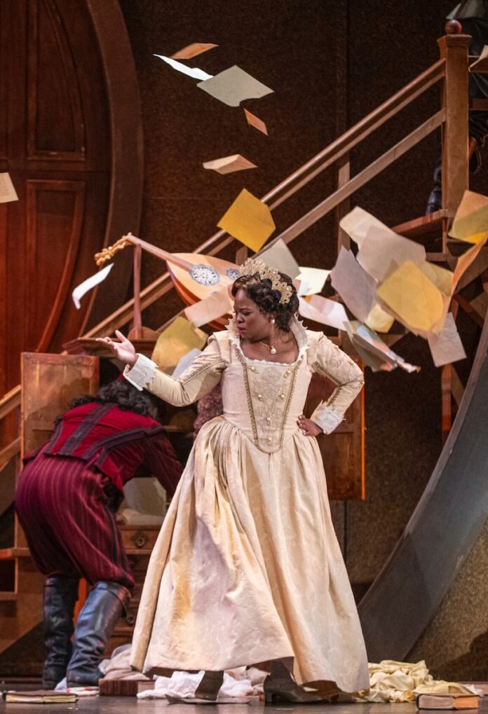 An opera singer poses in a long pink dress on stage with flyers flying around her.
