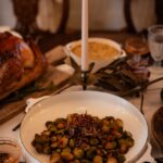 A white dish holds Roasted Brussels Sprouts with mushrooms and shallots, all sitting at a dinner table in front of a candlestick.