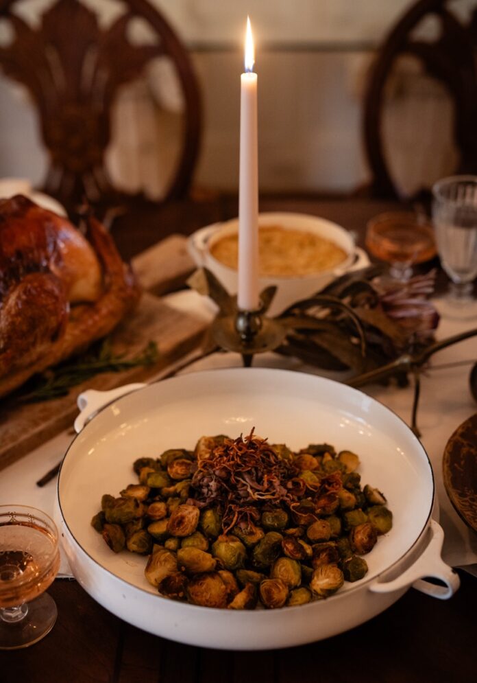 Roasted Brussels Sprouts with Mushrooms and Shallots A white dish holds Roasted Brussels Sprouts with mushrooms and shallots, all sitting at a dinner table in front of a candlestick.