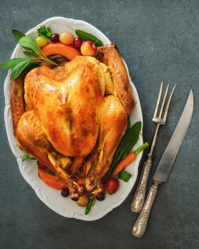A perfectly roasted whole chicken on a white platter garnished with grapes, apples, cranberries, and fresh sage, alongside a vintage carving set.