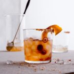 A glass of Rye Cranhattan on a table with ice, garnished with a cranberry and an orange peel.
