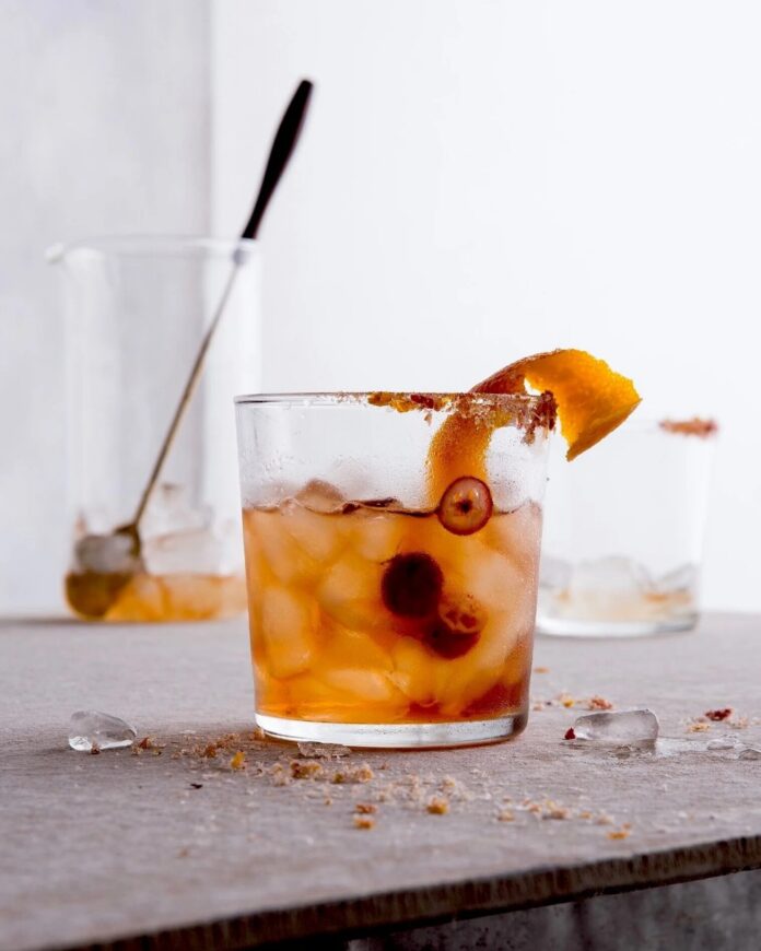 A glass of Rye Cranhattan on a table with ice, garnished with a cranberry and an orange peel.