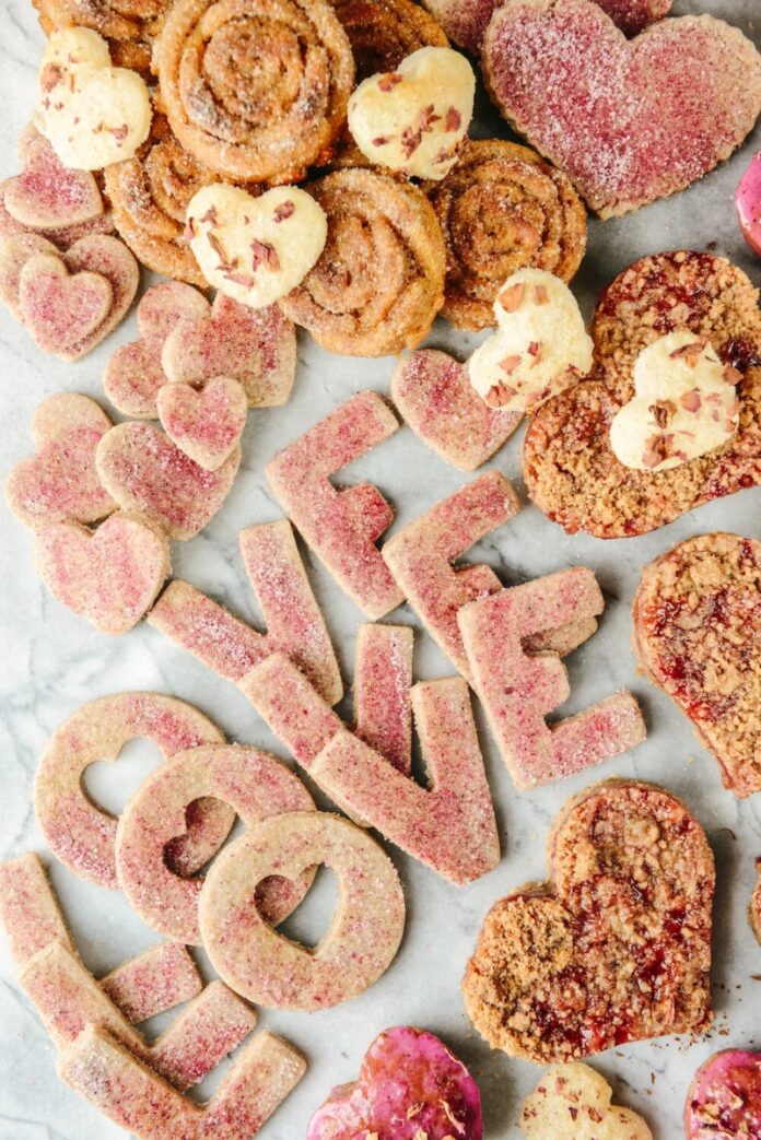 Heart-shaped and round cookies, some with a pink sugar dusting, arranged on a marble surface to spell out the word "LOVE" with individual letters.