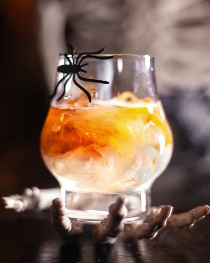 A clear glass of a creamy, orange-hued cocktail, garnished with a plastic spider, held aloft by a skeleton hand against a dark, foggy background.