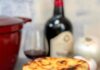 One Pot French Onion Soup A single serving of French onion soup in a red crock with melted cheese overflowing the sides, a bottle of red wine and a wine glass, and a large onion and garlic clove in the background.