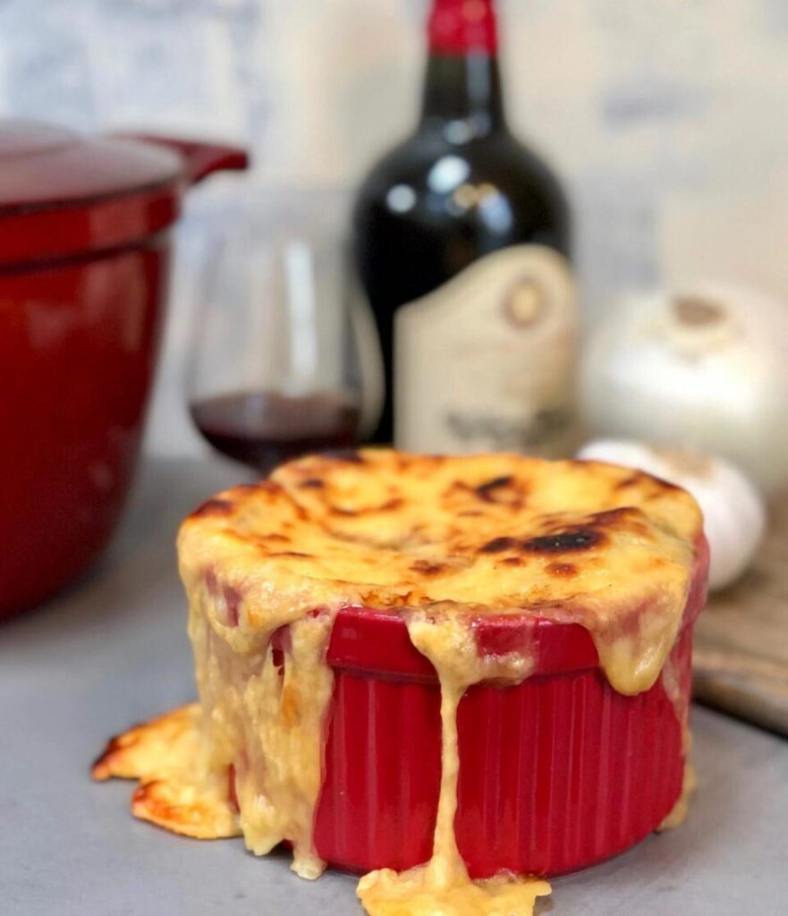 A single serving of French onion soup in a red crock with melted cheese overflowing the sides, a bottle of red wine and a wine glass, and a large onion and garlic clove in the background.