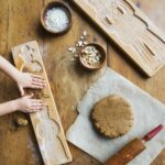 Two wooden gingerbread cookie molds, one of which a child's hands are pressing dough into, next to a ball of dough, a rolling pin, and small bowls of flour and sliced almonds.