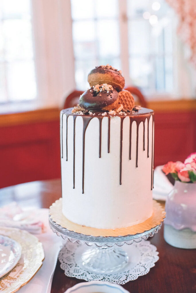 A tall white frosted cake with chocolate drip icing and two chocolate-covered donuts on top, on a glass cake stand.