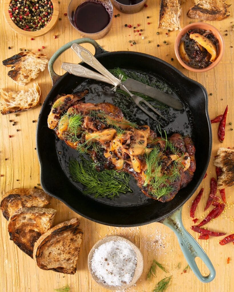 A large braised lamb shoulder in a cast iron pan, topped with dill and mushrooms, surrounded by toasted bread, chili peppers, and small bowls of salt and pepper.