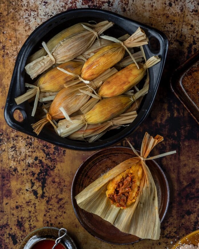 Traditional Tamales TABLE Magazine A black bowl filled with traditional tamales wrapped in corn husks, with one unwrapped tamale on a smaller plate showing a rich, red filling.