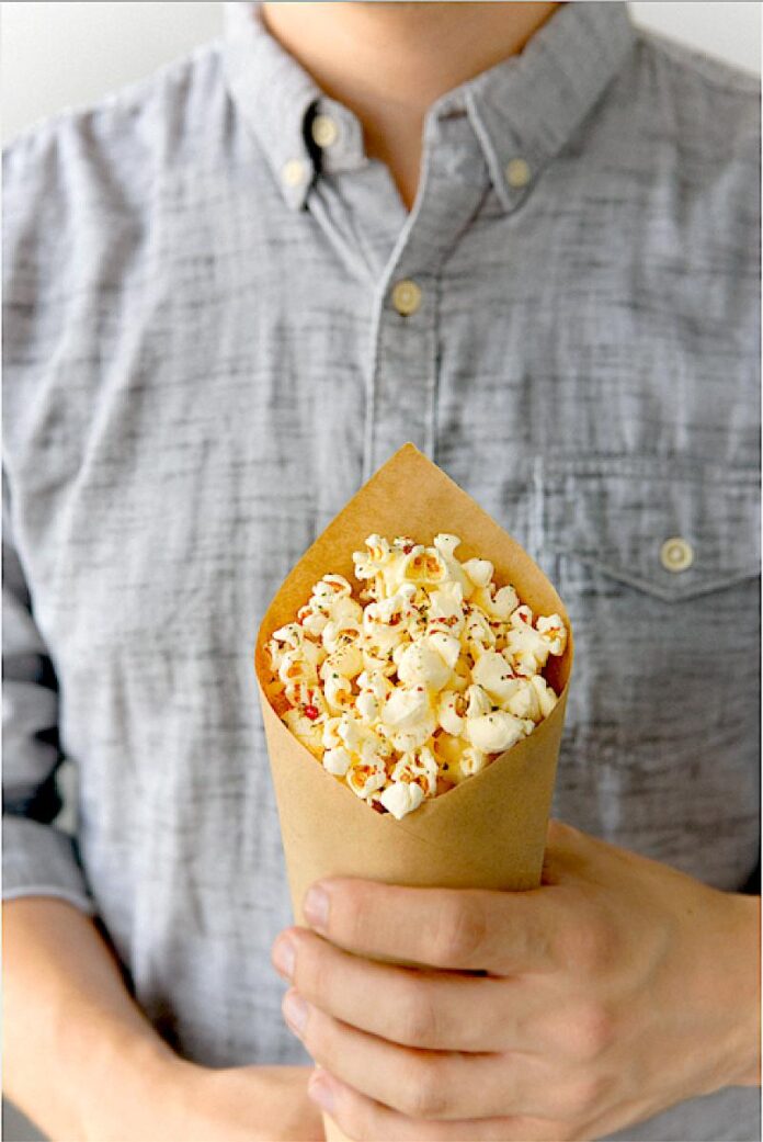 A person in a gray shirt holding a brown paper cone filled with popcorn, featuring a top-down view of the popcorn kernels.