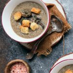 A bowl of vegan mushroom potato soup garnished with croutons and sliced mushrooms, with a ladle resting on a brown napkin.