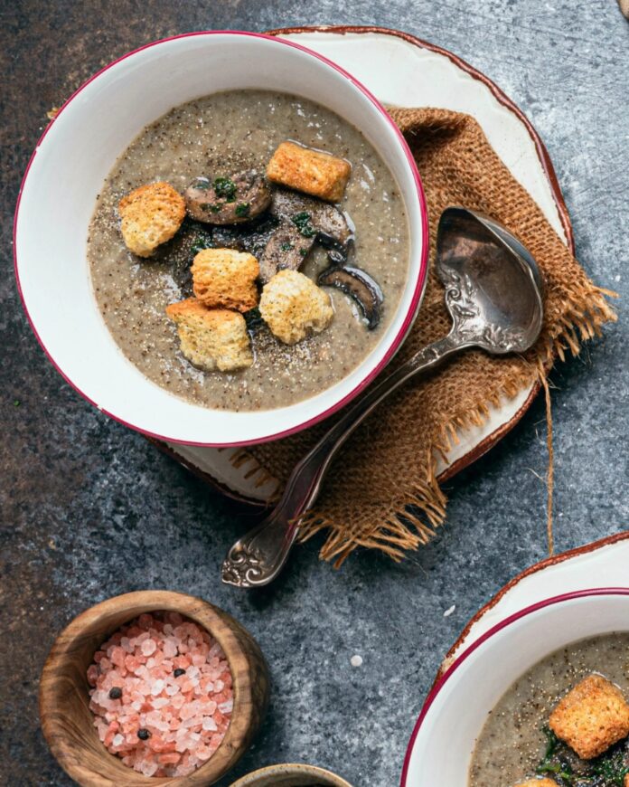 A bowl of vegan mushroom potato soup garnished with croutons and sliced mushrooms, with a ladle resting on a brown napkin.