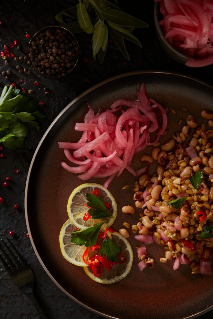A plate with a farro and black-eyed pea salad, topped with pickled red onions and garnished with lemon slices, parsley, and red chilies.