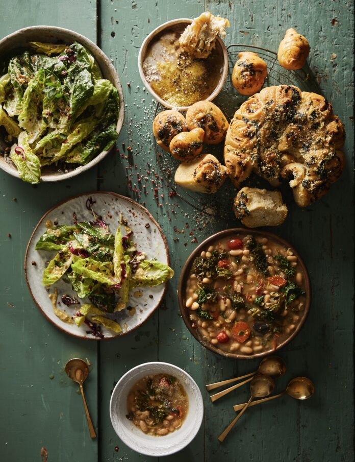 A green picnic table with a Caesar salad, garlic knots, and beans and greens bowl on top.