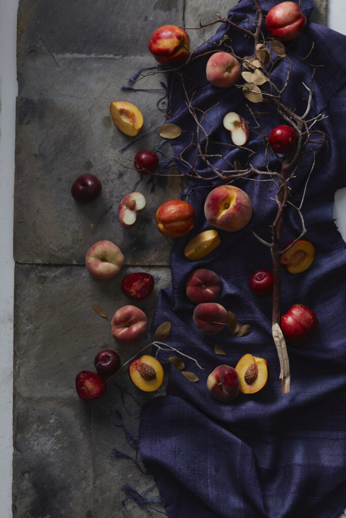 A bunch of peaches and plums from a local farm lay on a black table and royal blue cloth. 