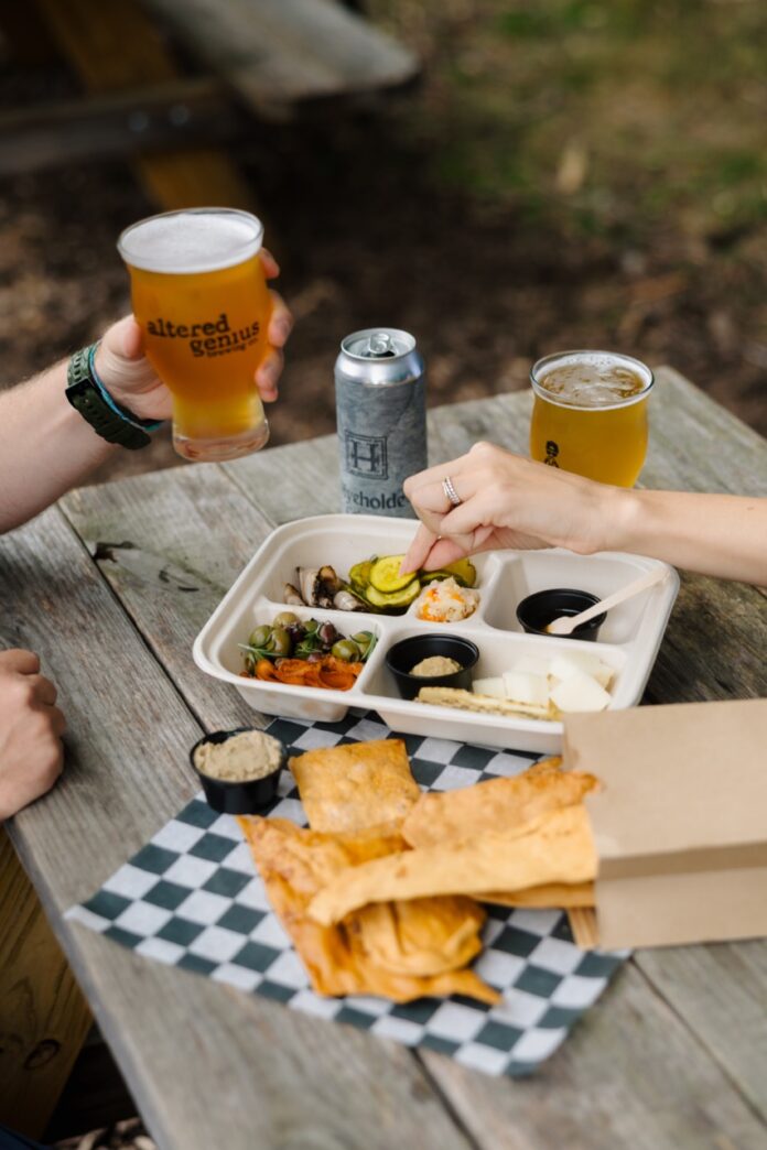 Two beers an a charcuterie board from Hyeholde and Altered Genius sit on a picnic table.