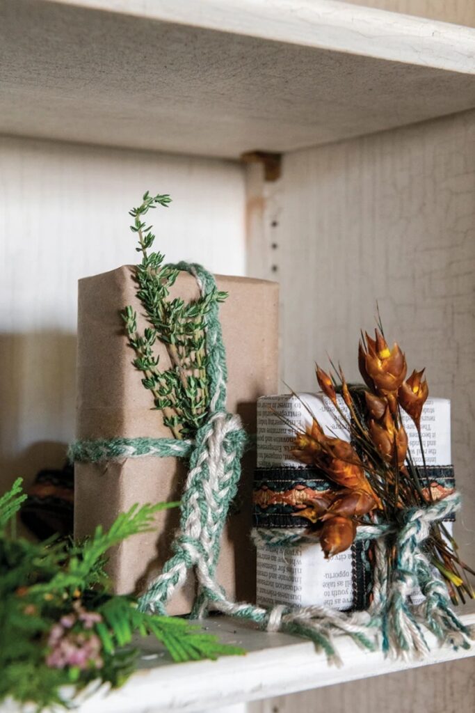 A gift wrapped in brown paper with a braided yarn ribbon and a sprig of fresh thyme, on a rustic shelf.