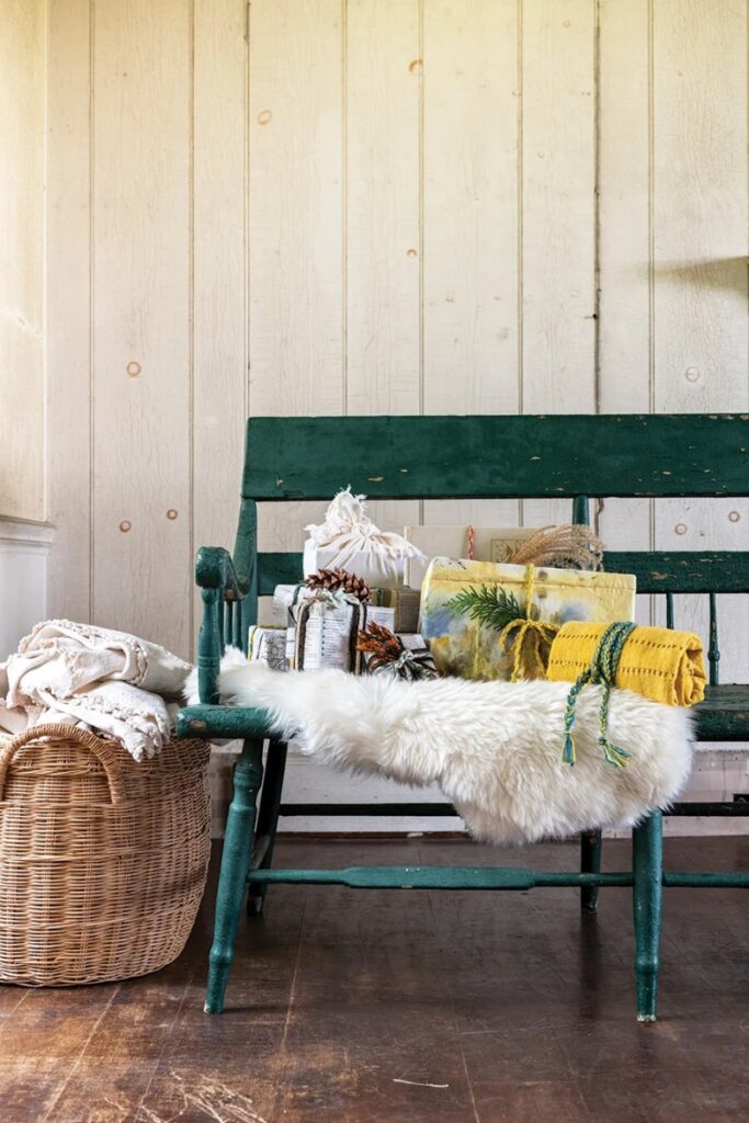 A vintage green wooden bench topped with a white fur throw, holding several gifts wrapped in reusable fabric and natural materials like furoshiki.