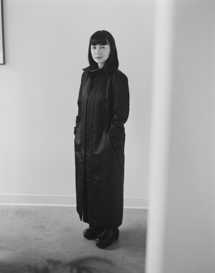A woman stands in a long black trench coat.