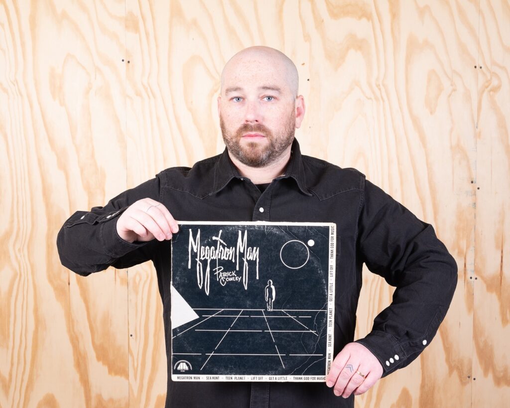 A man holds an art piece as he stands against a wood wall.