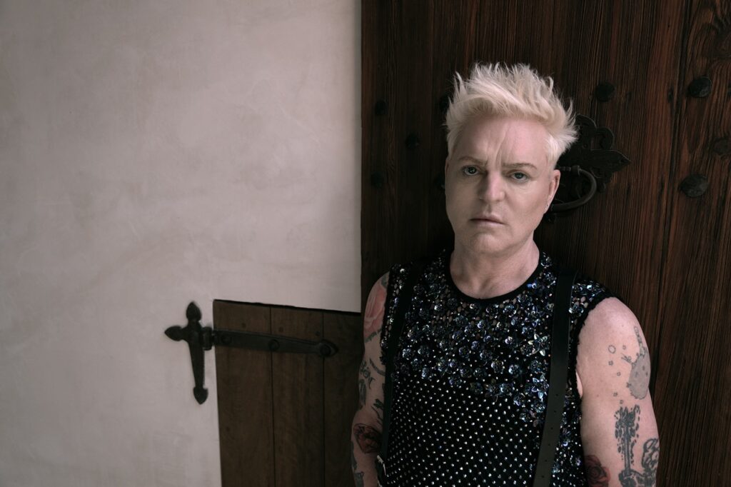 Andy Bell stands in a black sparkly tank top with white hair.