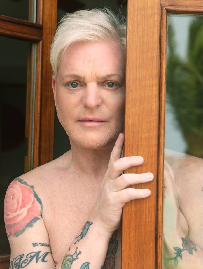 Erasure's Andy Bell on Solo Album 'Ten Crowns' and Touring A man stands behind a wood beam shirtless with white hair.