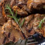 A close-up of grilled, sliced meatloaf topped with caramelized onions and brown gravy, garnished with fresh rosemary sprigs.