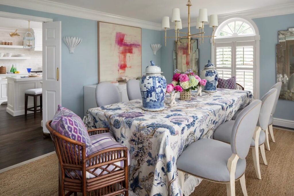 Classic dining room with light blue walls, a floral blue and white tablecloth, and ginger jars, opening into a bright white kitchen.