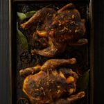 Two crispy roasted lemon chickens seasoned with sage, garnished with lemon zest and sage leaves on a dark baking sheet.