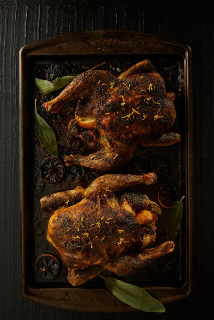 Two crispy roasted lemon chickens seasoned with sage, garnished with lemon zest and sage leaves on a dark baking sheet.