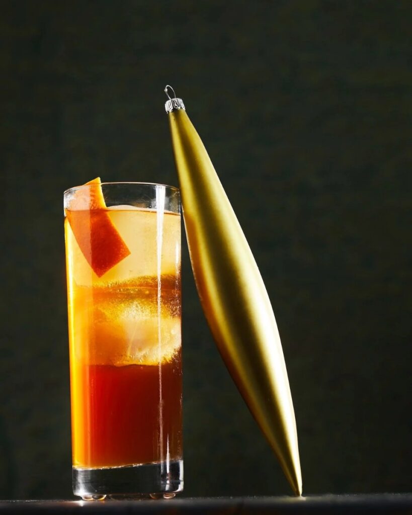 A rocks glass of drunk uncle cocktail with a large ice cube and an orange peel, next to a gold Christmas ornament.