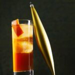 A rocks glass of drunk uncle cocktail with a large ice cube and an orange peel, next to a gold Christmas ornament.