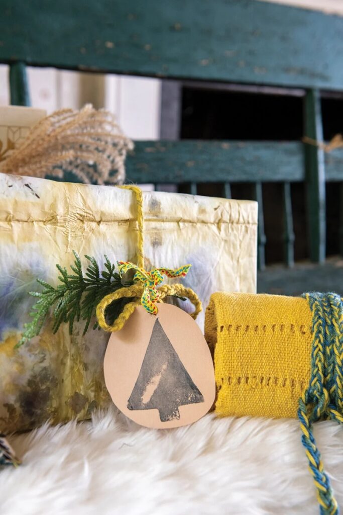 An eco-friendly wrapped gift, tied with twine and a natural paper tag with a stamped Christmas tree, next to a woven yellow fabric roll.