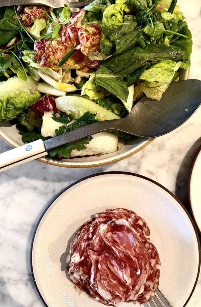 A bowl of salad above a plate of salami in a rose shape.