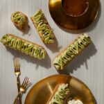 Four long tarts with green filling on a golden plate, with a coffee cup and two forks next to them on a white striped surface.