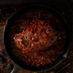 A cast iron pan filled with wild boar ragu, with various spices and peppers scattered on a dark surface around the pan.