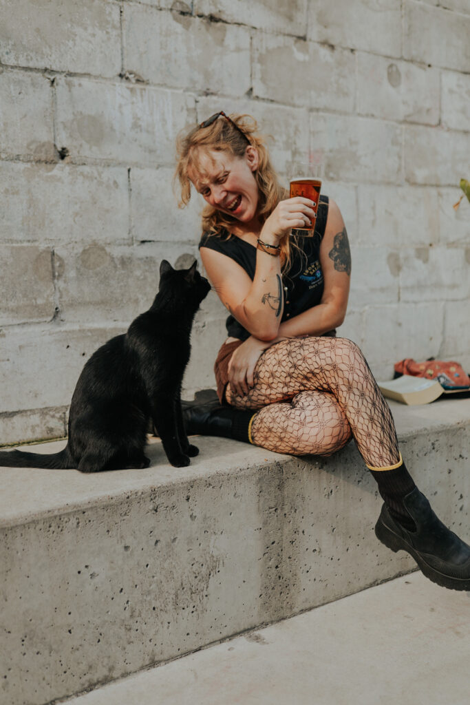 A girl sits outside in Pittsburgh with a beer in hand next to a black cat.