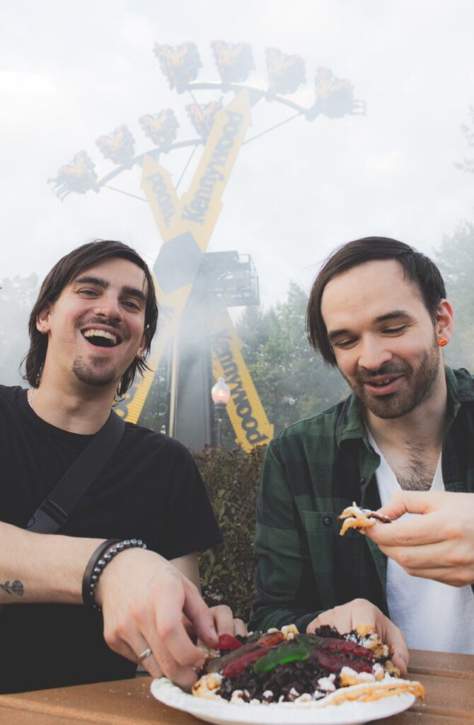 Two men share a funnel cake in front of the Arrow 360 ride.