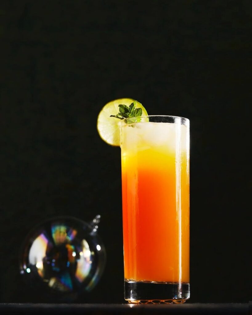 A tall glass of an orange and yellow kid's choice cocktail, garnished with a lime slice and mint, sits on a dark surface.
