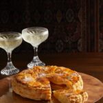 King Cake served on a round, flat, brownish plate with a piece cut and protruded out. Two stylish glasses filled with a drink.