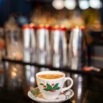 A holiday-themed white teacup with a holly and berry pattern, holding a brown cocktail garnished with dehydrated apple and allspice sugar, on a dark bar with shakers in the blurry background.