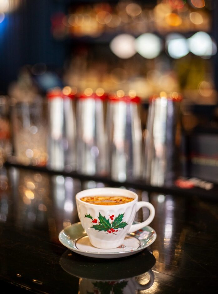 Kringle Cup TABLE Magazine A holiday-themed white teacup with a holly and berry pattern, holding a brown cocktail garnished with dehydrated apple and allspice sugar, on a dark bar with shakers in the blurry background.