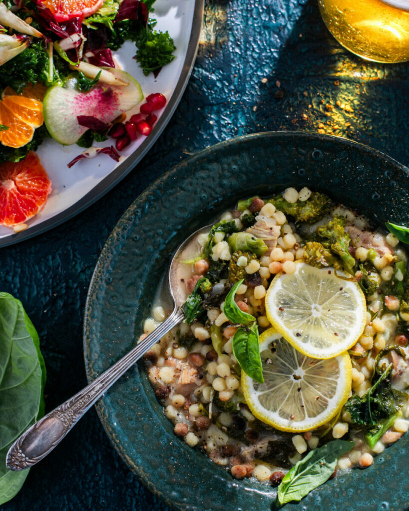 A bowl of soup with little round noodle balls, greens, chicken, and lemon slices on top sits in a dark teal bowl with a winter citrus salad sitting nearby.