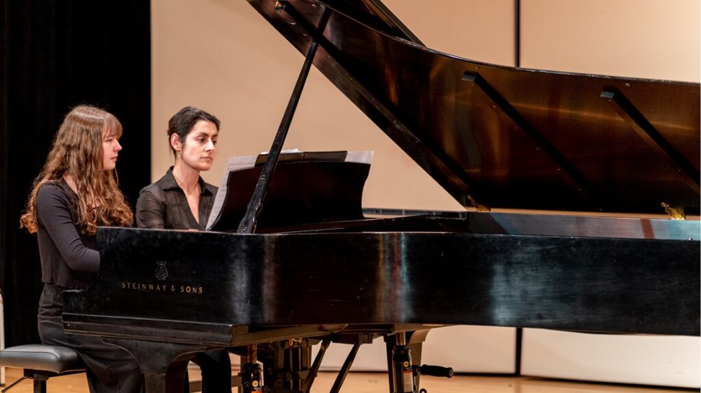 Two women sit at a black classic piano on a stage.