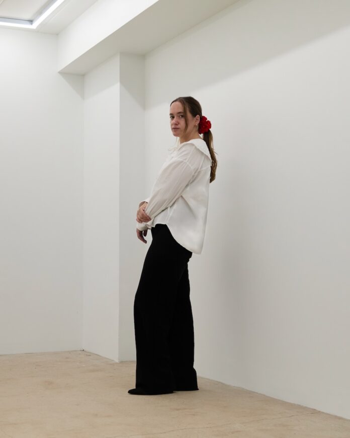 The owner of Romance Art Gallery stands against a white wall in a white shirt and black dress pants.